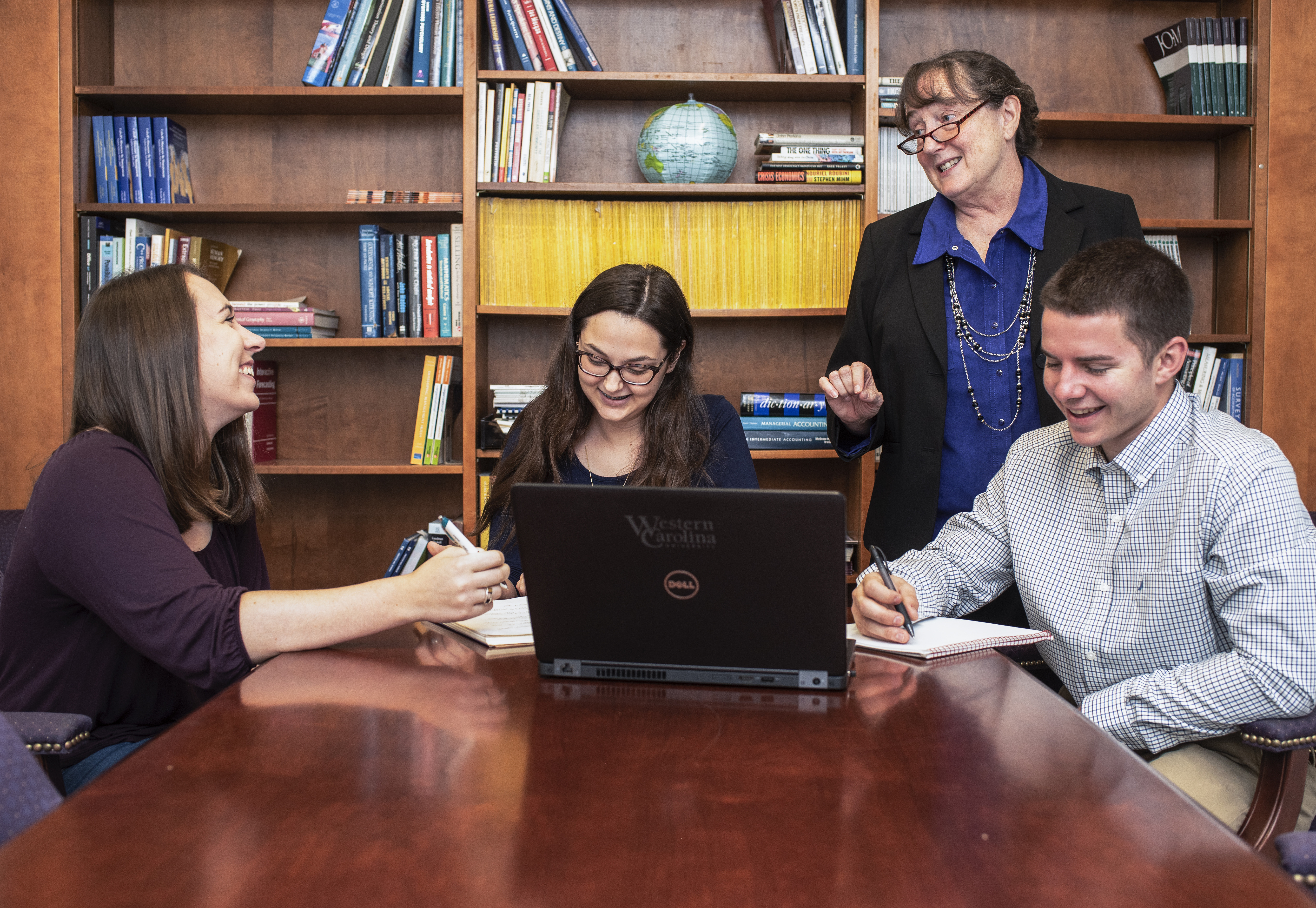 Hollye Moss stands and smiles while three students sit at a table working on laptops looking up at her and smiling