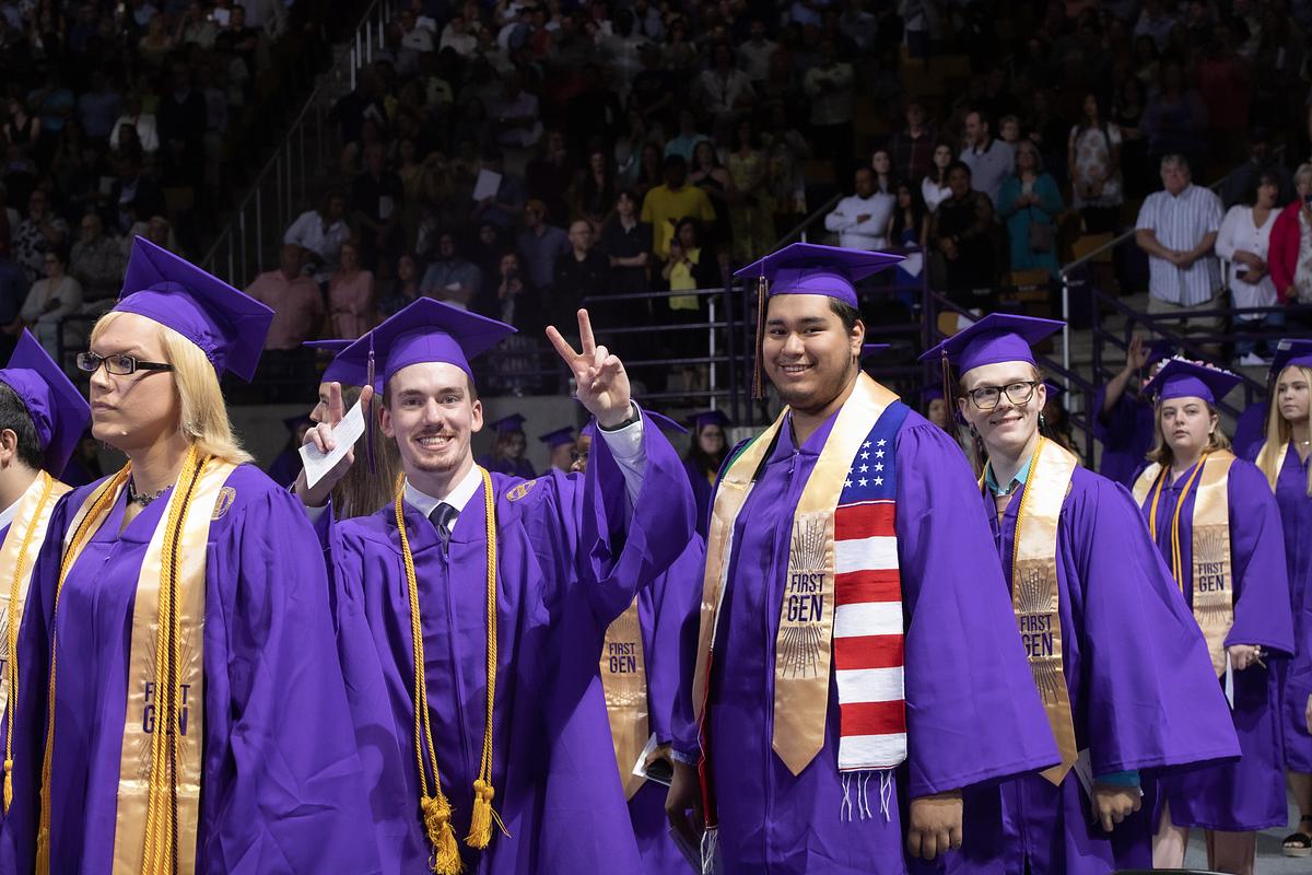 Students in purple gowns and gold First Gen stoles in line to walk across the stage, one student holding up peace sign