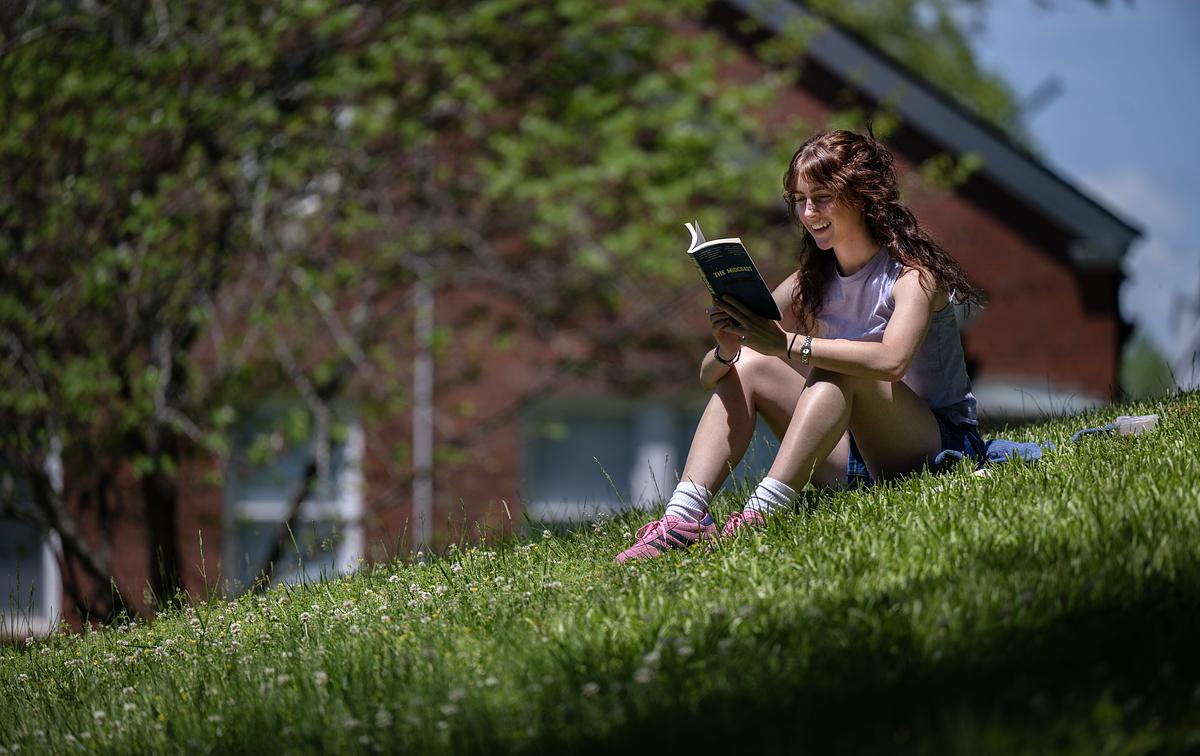 Young girl with curly hair reading a book while sitting on a grassy slope near an ivy-covered brick building.