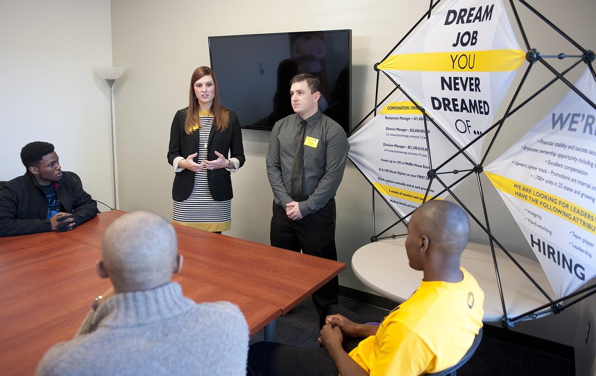 Two recruiters standing at the front of a room with a big company display with students sitting around a conference table